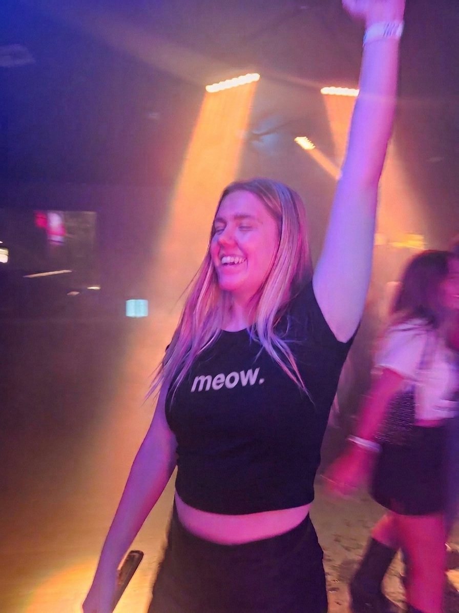 Woman dancing with one arm up, wearing a black Sparkbite baby tee that reads "meow."